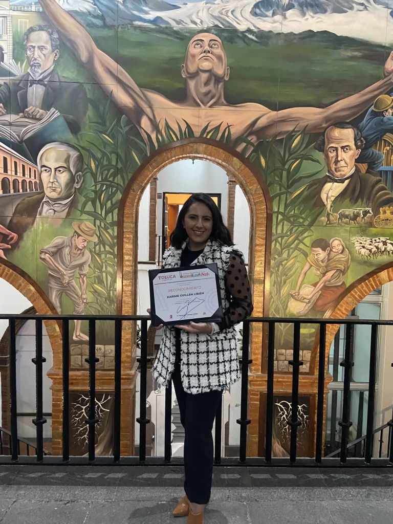 Karime Guillen sosteniendo un diploma frente a un mural histórico